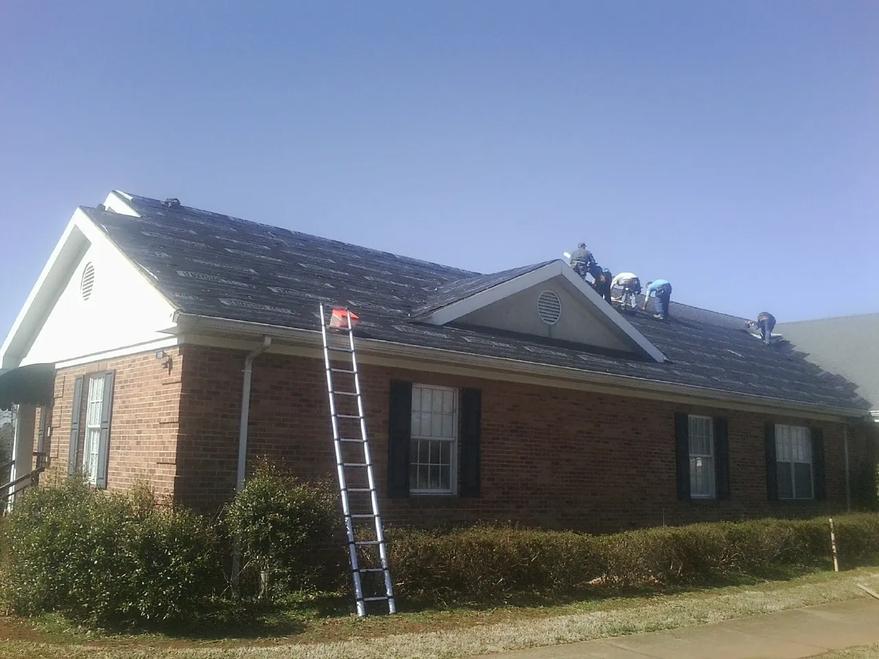Crew tearing shingles off a brick ranch roof with a ladder set — Emergency Roof Repair work by our crew in Sinai