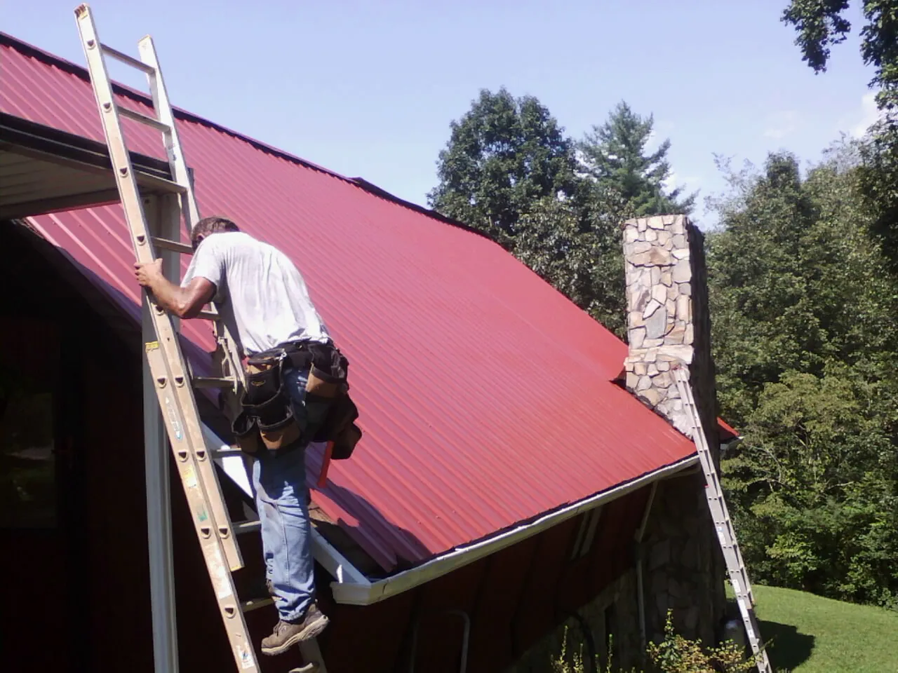 Roofer on a ladder beside a red metal roof and stone chimney — Soffit & Fascia Repair by Roofing Brothers in Sinai